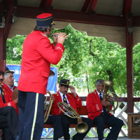 A band in vibrant red uniforms performs at an outdoor pavilion in Romania, with a focus on a standing trumpet player while his seated bandmates play brass instruments in the background, surrounded by lush greenery