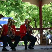 A brass band in red uniforms performs on an outdoor gazebo stage in Romania, with a tuba and horns in the foreground and a drummer at the back, all concentrating on their music.