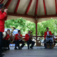 A brass band clad in traditional red and black uniforms performs enthusiastically under a gazebo in a lush, green park in Romania, with the conductor in front leading the ensemble.