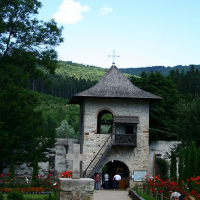 A rustic stone gateway with a wooden shingle roof leads into a serene courtyard surrounded by lush greenery in Romania, with visitors enjoying the tranquil surroundings.