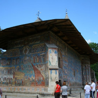 Visitors admire a historic painted monastery in Romania with intricate frescoes on its exterior walls, showcasing religious scenes under a clear blue sky.