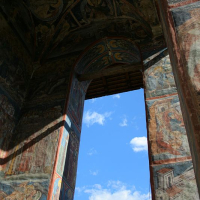 An ancient doorway adorned with intricate frescoes depicting religious scenes in Romania, opening to a bright blue sky, showcasing a blend of cultural history and natural beauty.