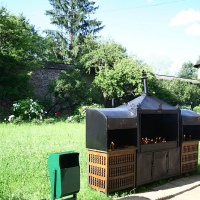 A large outdoor metal grill filled with lit charcoal is set in a verdant field with a green waste bin alongside, against a backdrop of an old stone wall and a medieval tower in Romania, surrounded by