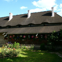 A rustic house in Romania with a distinctive thatched roof, featuring dormer windows, adorned with vibrant hanging flower baskets, and surrounded by a well-kept garden with blooming roses under a clear blue