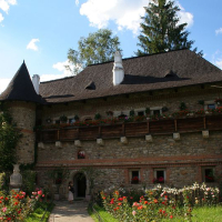 A picturesque stone-built castle in Romania with a conical turret, featuring wooden balconies adorned with flowers, surrounded by lush greenery under a clear blue sky.