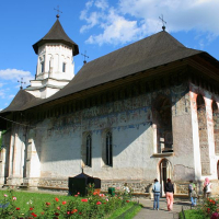 A historic church in Romania with a distinct stone bell tower and frescoed exterior walls, surrounded by lush greenery and flowering plants under a clear blue sky, while visitors approach its entrance.