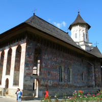An ornate medieval monastery in Romania with vibrant frescoes on its exterior walls, under a clear blue sky, is admired by visitors strolling through its surrounding garden with blooming flowers.