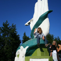 A person sits on a vibrant, oversized sculpture of a green and white hand in Romania, basking in the sunlight against a clear blue sky, surrounded by tall green trees.