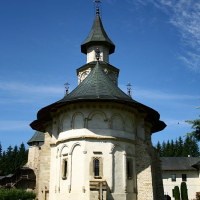 A historical church in Romania with a distinctive wood-clad spire and stone base stands gracefully under a clear blue sky, surrounded by manicured lawns and lush greenery, epitomizing architectural elegance