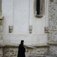 A solitary clergyman in traditional black vestments carries a blueprint while walking on the grass beside an old stone church in Romania, reflecting a blend of history and ongoing care or restoration.