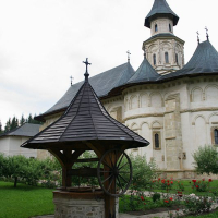 A traditional wooden well stands in the foreground of an ornate church with a prominent spire in Romania, surrounded by lush greenery and red flowers, evoking a serene, pastoral atmosphere.