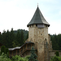 An ancient stone tower with a conical roof stands guard beside a manicured pathway lined with vibrant flowers in Romania, with lush green trees under a cloudy sky in the background, evoking a sense of
