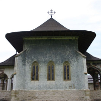 An ancient stone church with a large conical roof, featuring elongated windows and a cross atop, nestled in a serene grassy field in Romania.