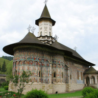 An historical monastery adorned with frescoes in Romania, nestled in a lush, green valley under a cloudy sky, showcases a traditional spired tower and a wrap-around porch with religious murals.