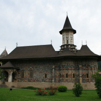 A traditional monastery in Romania with elaborate exterior frescoes and a prominent bell tower, surrounded by lush greenery under a cloudy sky.