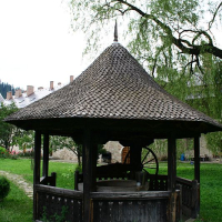 A traditional Romanian wooden well gazebo with a shingled roof, nestled in a lush green meadow with trees and a backdrop of white buildings with red roofs.