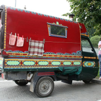 A woman stands beside a vibrantly decorated, red caravan with cultural Romanian patterns, parked in a roadside area, suggesting a glimpse into a nomadic or traveler lifestyle.
