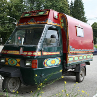 A woman talks on a mobile phone beside a colorfully painted, three-wheeled vehicle, resembling a decorated auto-rickshaw with an enclosed cargo area, parked on a sunny street in Romania with