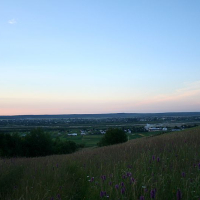 A serene dusk falls over the rural landscape of Romania, with the gentle slope of a flowering meadow in the foreground descending to a tapestry of fields and houses, all under the watchful gaze of