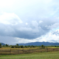 A serene rural landscape in Romania under a dramatic sky, with lush greenery, a rustic wooden fence leading the eye towards distant mountains, and heavy clouds suggesting an approaching storm.