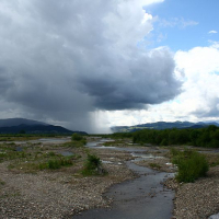 A vast, serene landscape in Romania with a meandering river amidst pebbled banks under a dramatic sky, where a localized rainstorm pours from a dark cloud onto distant hills, contrasting with the sun