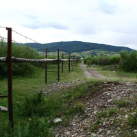 A rustic wooden footbridge with metal railings crosses over a dry rocky creek bed in Romania, leading towards a lush green valley with rolling hills under an overcast sky.