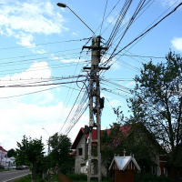 An electric pole stands at a rural street's edge in Romania, its arms entangled with a complex web of wires under a cloudy sky, flanked by greenery and quaint houses with picket fences