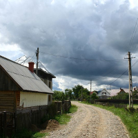 A rustic dirt road leads through a tranquil village in Romania with wooden houses under a dramatic sky, hinting at an approaching storm.