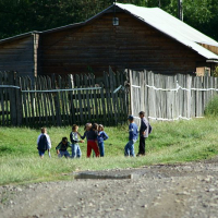 A group of children walk beside a wooden fence in a rural Romanian setting, with wooden houses in the background and a gravel road in the foreground, suggesting a communal or village lifestyle.