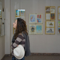 A woman with curly hair, holding a white hat, contemplatively observes colorful artwork displayed in simple frames on a gallery wall in Romania, illuminated by soft lighting.