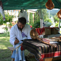 A craftsman in traditional Romanian attire meticulously carves wood under a canopy at an outdoor market, surrounded by artisanal creations, symbolizing cultural heritage and the enduring artistry of handcrafting.