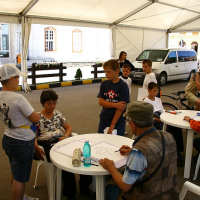 Children gather around a table at an outdoor workshop in Romania, engaging attentively with an instructor who is demonstrating or instructing, encouraging interactive learning in a casual, tented setting.