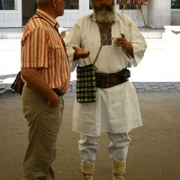Two men are standing and talking. The man on the left wears glasses, a short-sleeved orange-striped shirt, beige pants, and brown shoes. The man on the right, with a white beard, is dressed in traditional Romanian folk attire: a white shirt, pants, a dark hat, and traditional footwear.