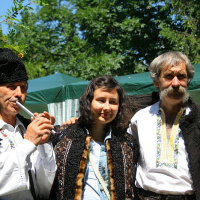 Three individuals in traditional Romanian attire—two men with vests and furry hats flank a smiling woman in a blouse—pose outdoors on a sunny day, with the elder man on the right holding a wooden flute