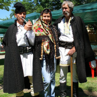 Three individuals in traditional Romanian attire pose outdoors; two men in embroidered vests and fur hats flank a woman in a colorful headscarf, exemplifying cultural heritage.
