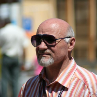 A man with a mustache wearing sunglasses and a striped shirt stands in focus, with a blurred background indicating a bustling outdoor setting in Romania.