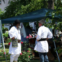 Two individuals in traditional Romanian folk costumes engage in a conversation at a sunny outdoor market, surrounded by tents and handicrafts.