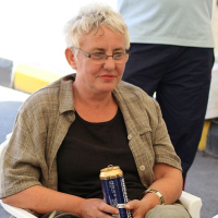 A woman with short, light-colored hair, wearing glasses and a checkered jacket, casually sits holding a can, attentively listening with a slight smile in a tented area in Romania.