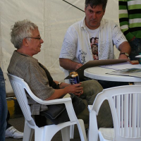 Two individuals engaged in conversation at a casual gathering in Romania, with one sitting across a table holding a beverage can, surrounded by a relaxed atmosphere under a tented structure.