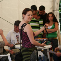 A woman stands smiling at the camera, holding a bag, in a tent in Romania where people are engaged in various activities, some sitting and others conversing, suggesting a casual gathering or event.