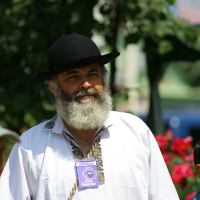 A man with a full beard and a black hat, wearing a traditional Romanian folk shirt adorned with a badge, gazes into the distance at an outdoor event.