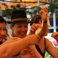 Three people joyfully stretch a piece of cheese, displaying its stringy texture, while sharing a moment of camaraderie around a table with food and flowers in the background in Romania.