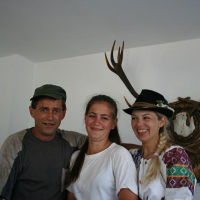 Three individuals pose smiling: a man in a cap, a young woman in white, and another with a traditional Romanian hat, with an antler display and clock in the background.