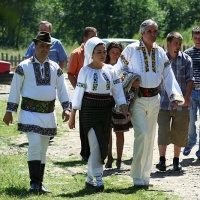 A group of people, some dressed in traditional Romanian embroidered outfits, walk together in the sunshine at a cultural event with nature and parked cars in the background.