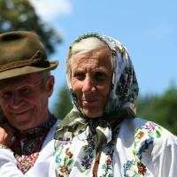 An elderly couple in traditional Romanian attire, with the woman wearing a floral headscarf and embroidered shirt, stands close together against a sunny, tree-lined backdrop, evoking a sense of cultural heritage and
