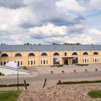 A large, yellow, historic building within the Daugavpils fortress boasts a long facade featuring arched windows under a gray roof. The cobblestone courtyard and landscaped shrubs lie in the foreground. The partly cloudy sky casts soft light, enhancing the tranquil atmosphere.