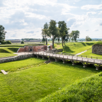 A scenic view of Daugavpils Fortress reveals a grassy landscape, a wooden bridge spanning a moat, and striking red-brick structures. Trees are scattered around the area, and the sky is partly cloudy, casting soft light over this historic Latvian scene.