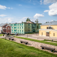 A scenic view of Daugavpils, with a cobblestone path in the foreground, lined by wooden benches on grassy slopes. To the left, a red brick building with arches and to the right, a yellow building with gray roof. In the background are greenish buildings under a blue sky with clouds.