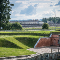 A serene landscape features the historic Daugavpils fortress in the foreground, its brick walls perched on a grassy slope. In the background, a tree stands near a road, while expansive industrial buildings are visible under Latvia's cloudy sky. Two people stand near the entrance of the fort.