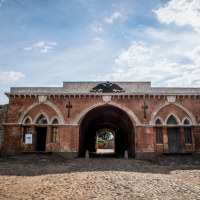 A historic fortress entrance in Daugavpils, Latvia, with arched doorways made of red brick and stone. The structure features decorative elements above the arches and framed windows. A cobblestone pathway leads to the entrance under a partly cloudy sky, with two flags visible on the right side.